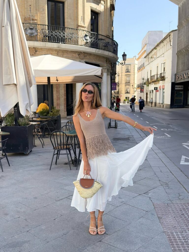Woman in a taupe fringe-hem knit tank and flowing white midi skirt with lace-up sandals, twirling the skirt on a street corner in Andalusia, Spain
