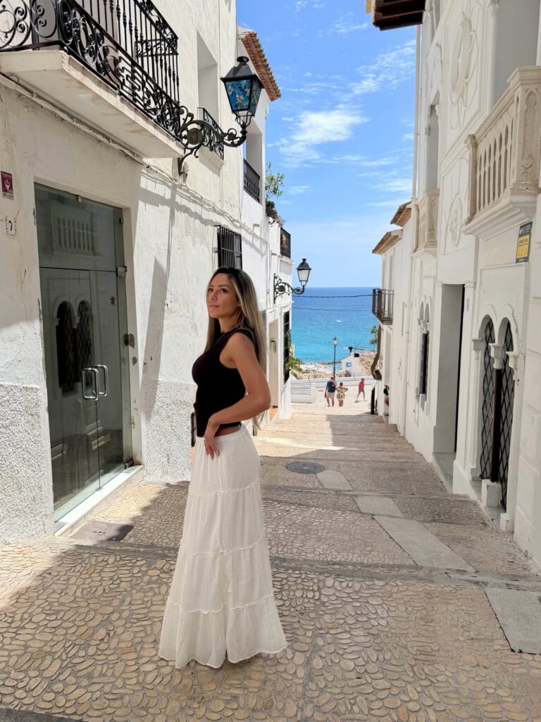Woman in a black tank top and white tiered maxi skirt looking over her shoulder on a whitewashed street with the Mediterranean Sea visible in Altea, Spain