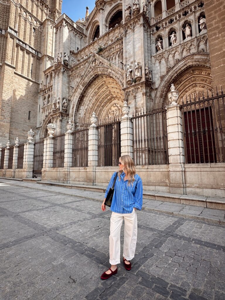 Woman in an oversized blue striped button-down shirt and cuffed white pants with burgundy velvet Mary Jane flats in front of Toledo Cathedral, Spain