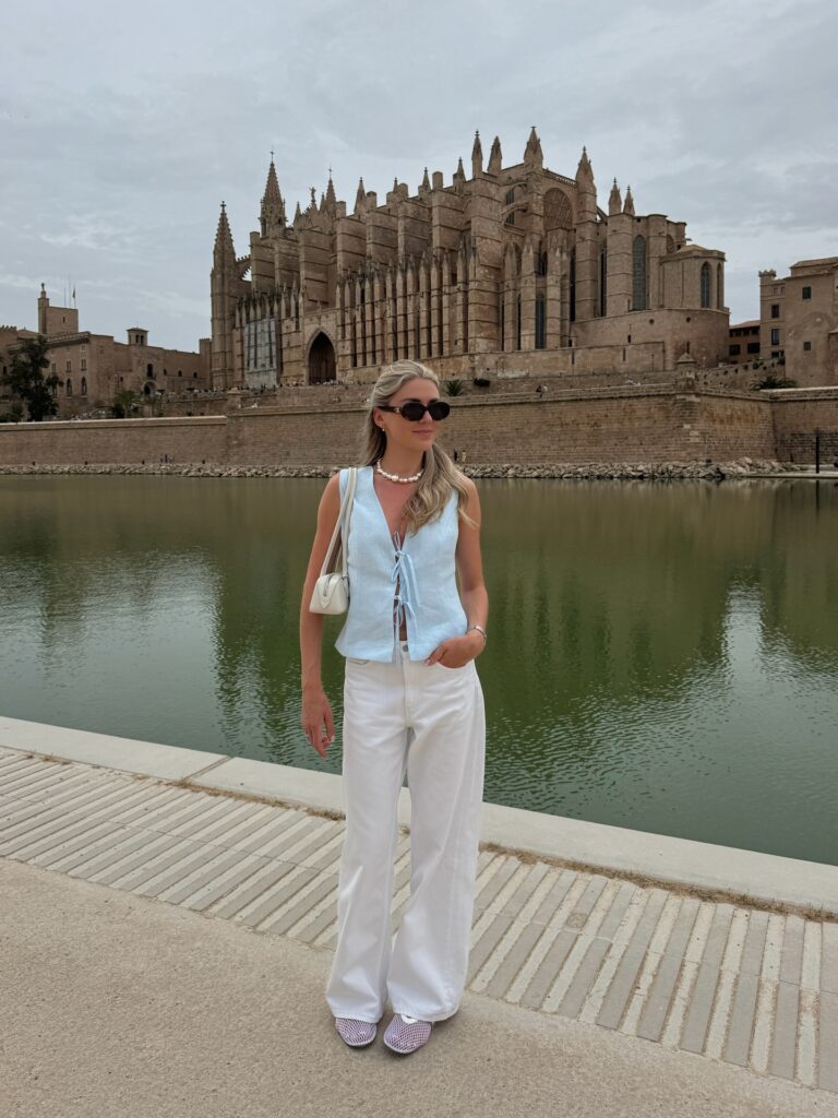 Woman in a light blue linen tie-front vest and white wide-leg jeans with a pearl necklace, standing in front of Palma Cathedral in Mallorca, Spain