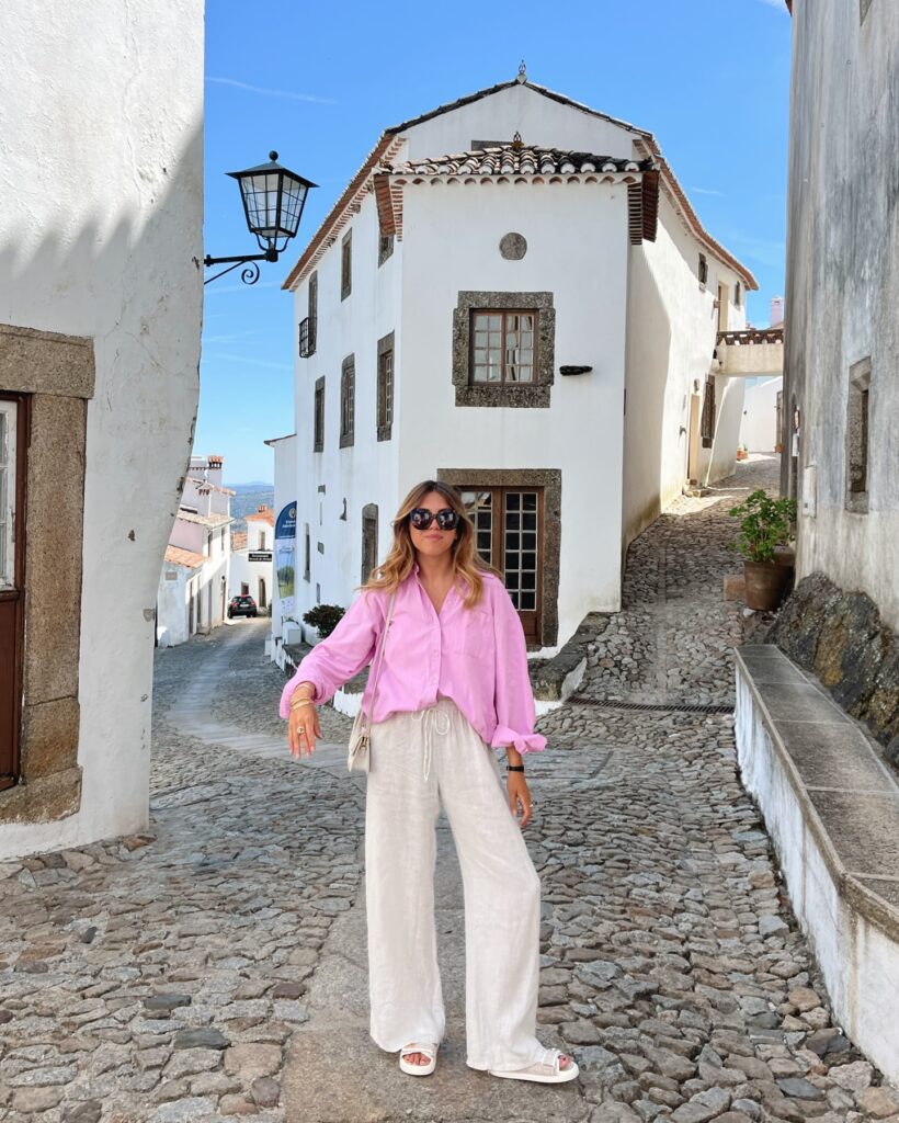 Woman in an oversized pink button-down shirt and beige linen drawstring pants with slide sandals on a cobbled street in a whitewashed hilltop village