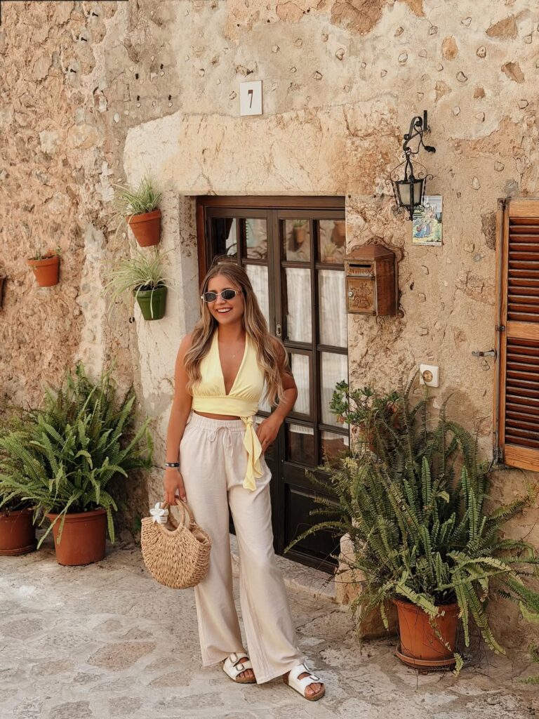 Smiling woman in a yellow halter wrap top and beige linen pants with white Birkenstock sandals and a round straw bag outside a stone house in Valldemossa, Mallorca