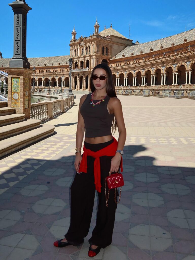 Woman in a brown tank top and black wide-leg cargo pants with a red sweater tied at the waist and a red mini bag at Plaza de España in Seville, Spain