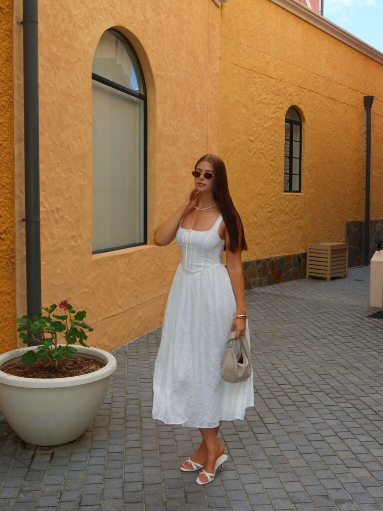 Woman in a white corset-bodice midi dress with oval sunglasses and a woven bag standing beside an orange stucco wall
