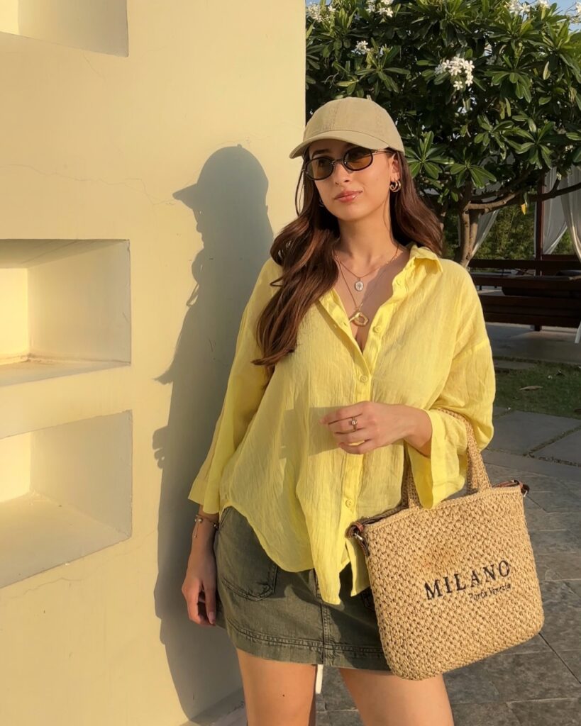 Woman in a sheer yellow oversized shirt, olive denim mini skirt, beige baseball cap, and woven tote in golden evening light