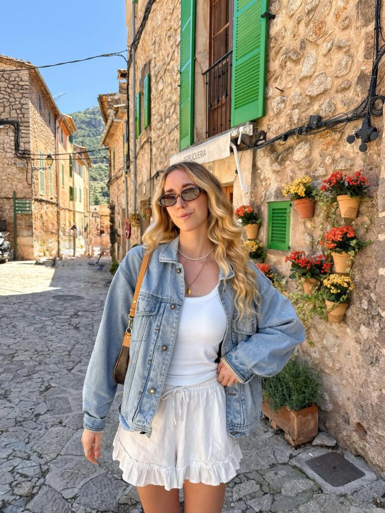 Woman in an oversized light-wash denim jacket, white tank, and white ruffled shorts on a stone street with green shutters and flower pots in Valldemossa, Mallorca