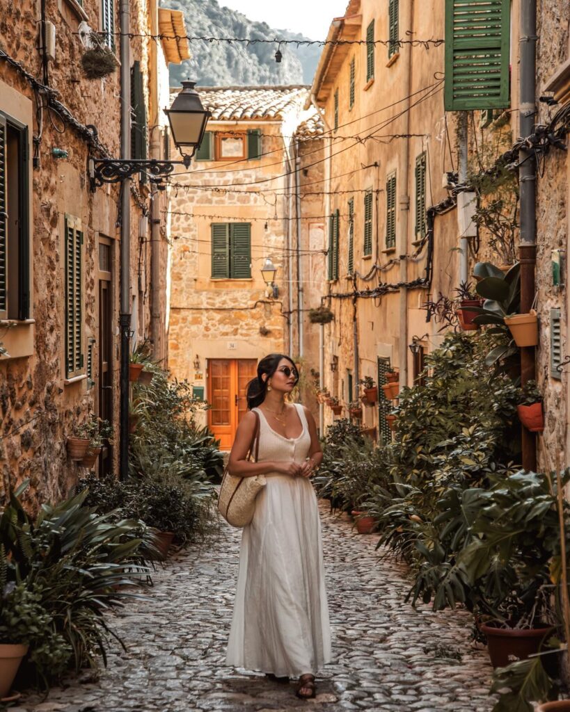 Woman in a white knit-bodice maxi dress with a straw shoulder bag walking a plant-lined cobblestone lane in Valldemossa, Mallorca