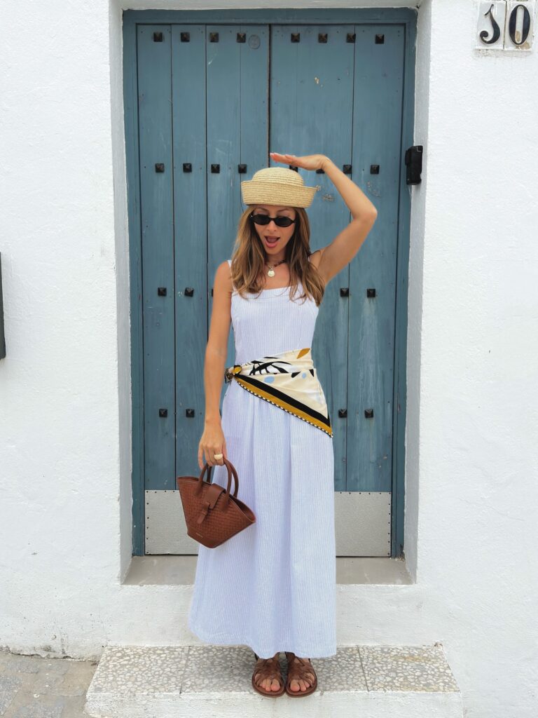 Woman in a blue and white striped maxi dress with a printed silk scarf tied at the hip and a straw hat, standing in front of a blue door in a whitewashed Spanish village