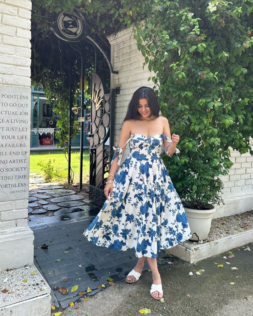 Woman in an off-the-shoulder white midi dress with blue floral print and white slide sandals standing by a garden gate