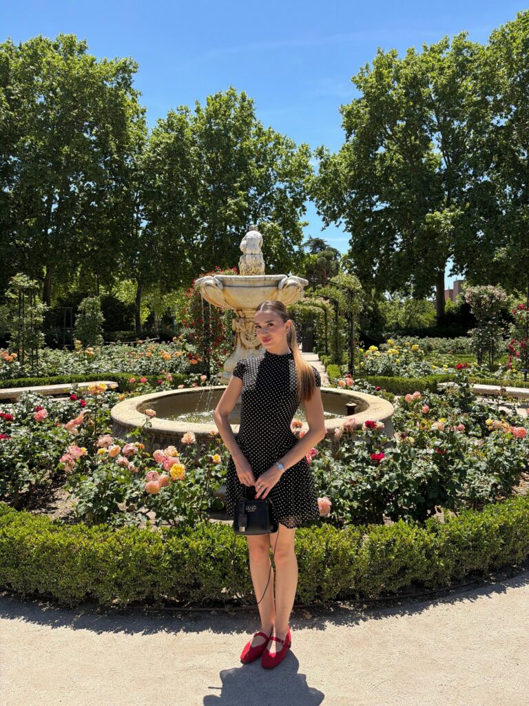Woman in a black polka dot mini dress with red Mary Jane flats standing by a rose garden fountain in Madrid's Retiro Park