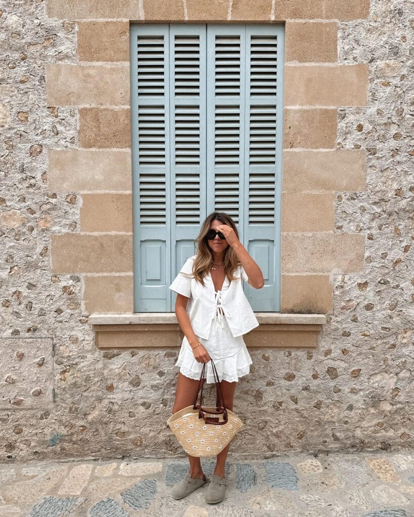 Woman in a white tie-front linen top and matching ruffled skort with suede clogs and a daisy-embroidered basket bag, standing under pale blue shutters on a stone wall in Mallorca