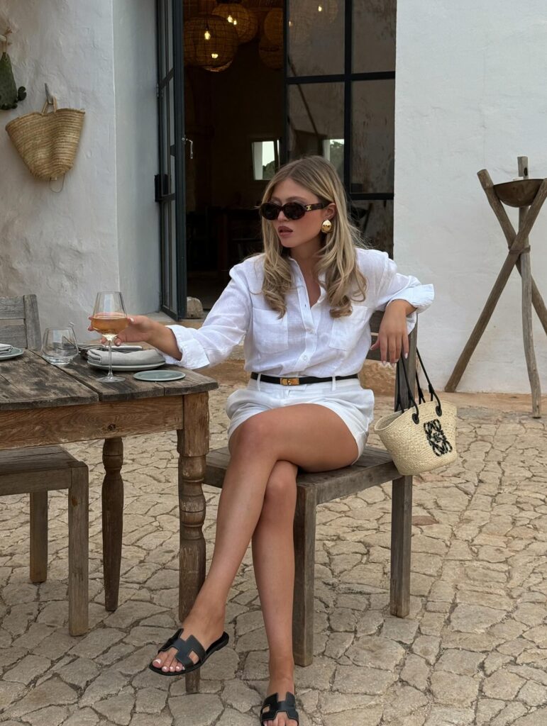 Woman in an oversized white linen shirt and white shorts with a slim black belt, black slide sandals, and a basket bag, seated at a rustic stone-courtyard table with a glass of wine