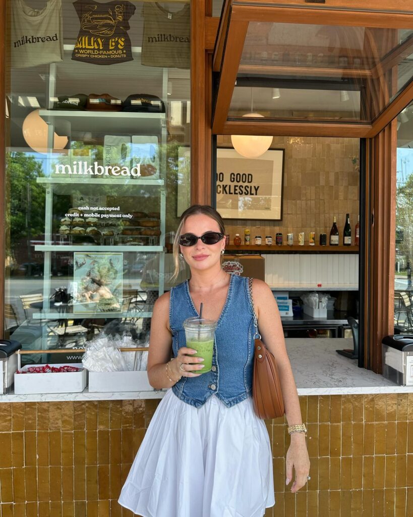 Woman wearing a denim vest and full white poplin mini skirt holding an iced matcha at a bakery walk-up window
