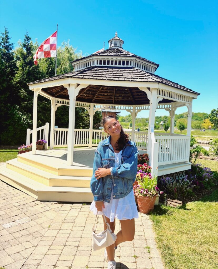 Woman wearing an oversized denim jacket over a white ruffled mini dress standing in front of a white gazebo surrounded by flowers