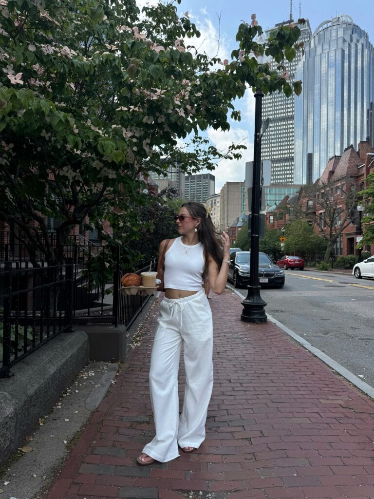 Woman wearing a white cropped tank top and white linen drawstring pants holding an iced coffee and croissant on a brick sidewalk