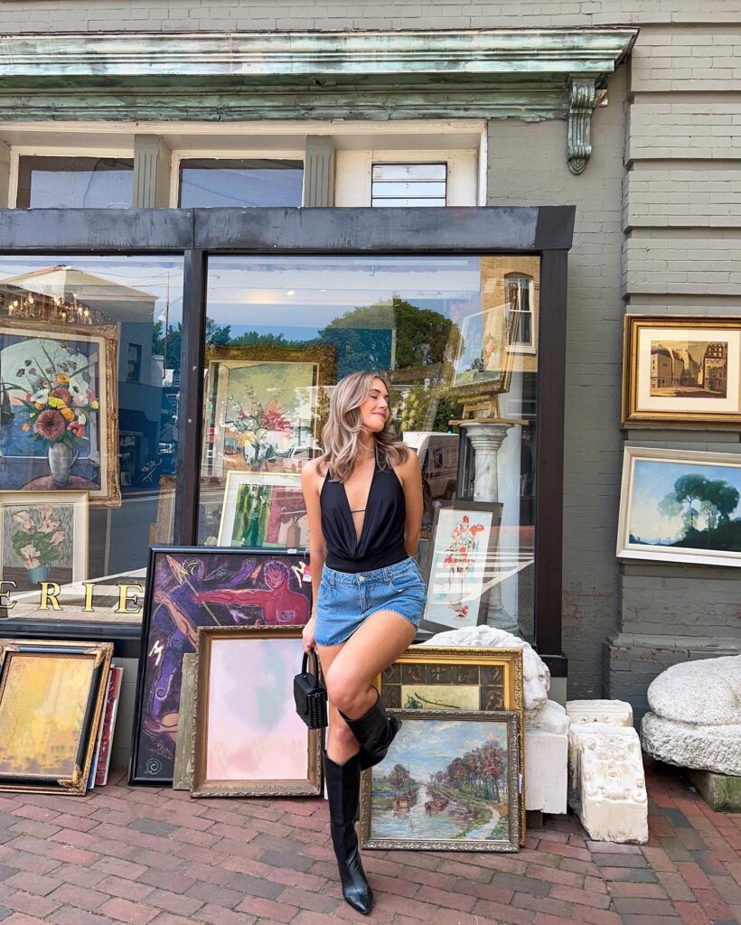 Woman wearing a black halter bodysuit, denim mini skirt, and black knee-high boots standing in front of an art and antique shop window