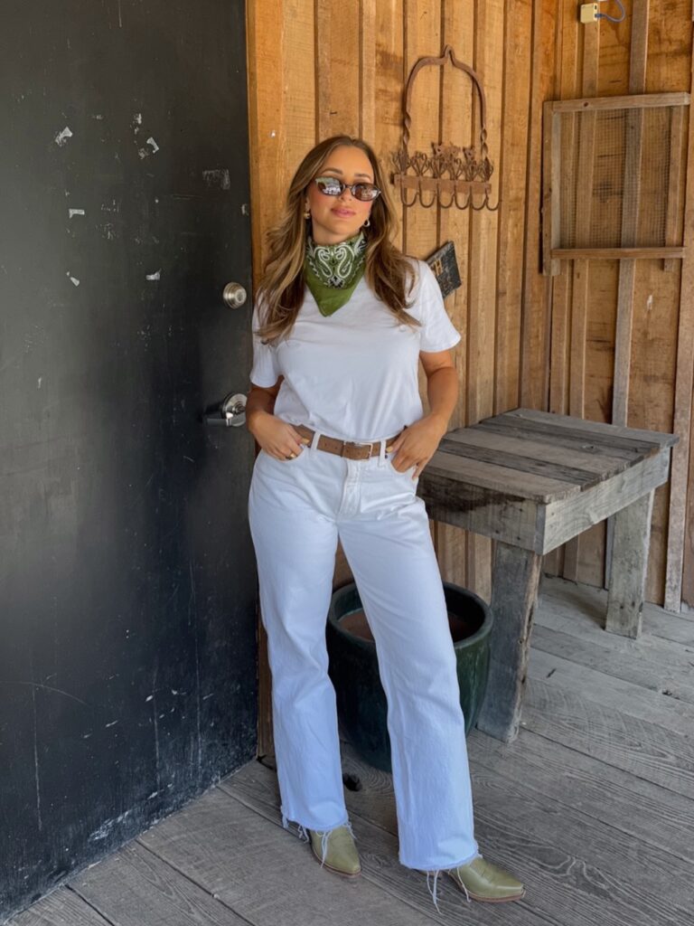 Woman wearing a white t-shirt, white jeans, a green bandana tied at the neck, and olive green cowboy boots on a rustic wooden porch