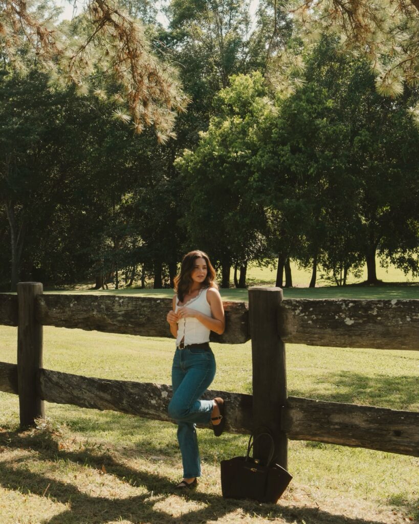 Woman wearing a white lace-trim tank top and straight-leg jeans with mary jane flats leaning against a rustic split-rail fence