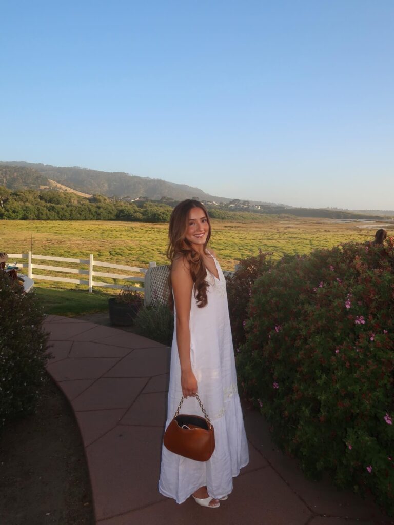 Woman wearing a flowy white maxi dress with white heeled sandals and a brown shoulder bag on a garden path at golden hour