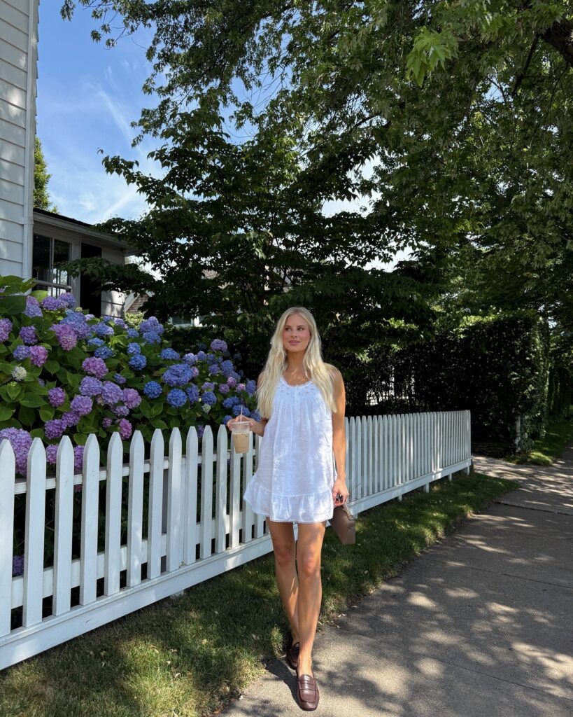 Woman wearing a white sleeveless mini dress with brown loafers walking past a white picket fence lined with hydrangeas, holding an iced coffee