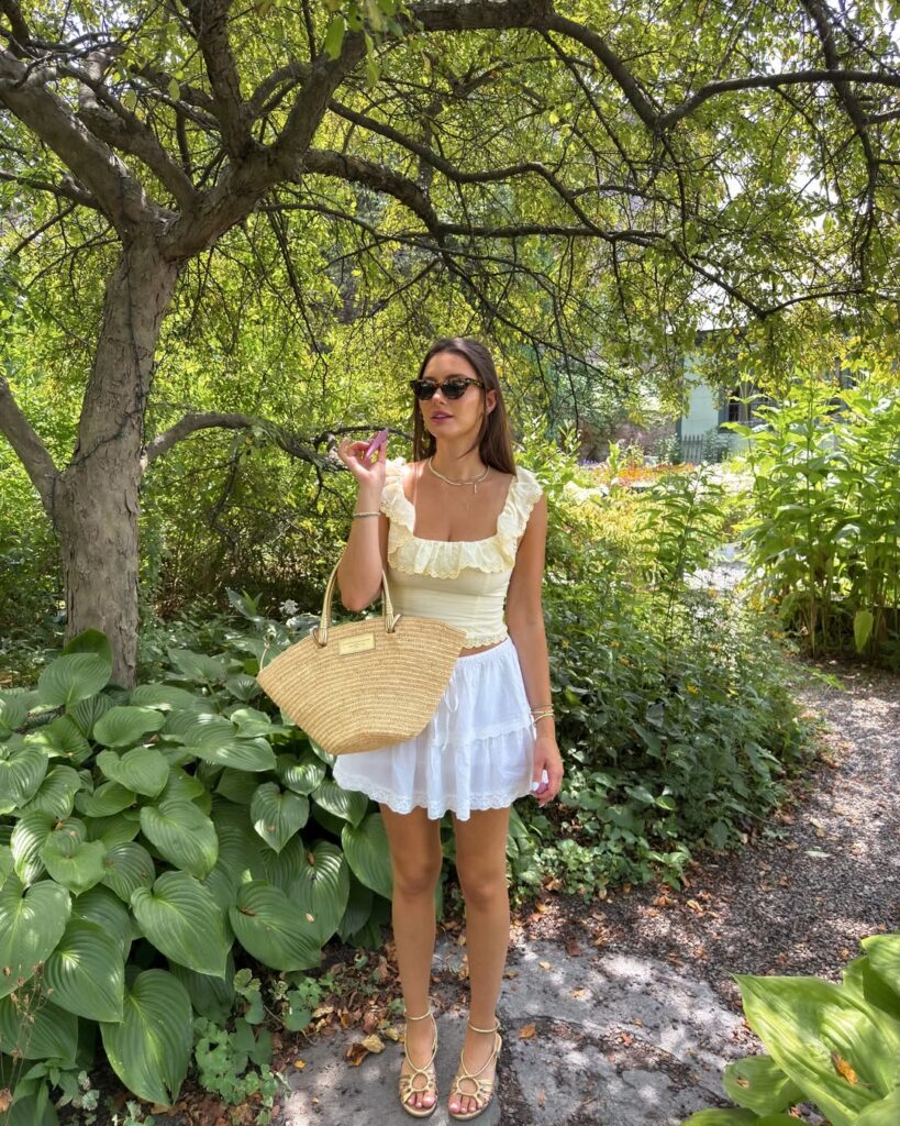 Woman wearing a pale yellow ruffle-trim top and white tiered mini skirt with gold sandals and a straw tote in a leafy garden