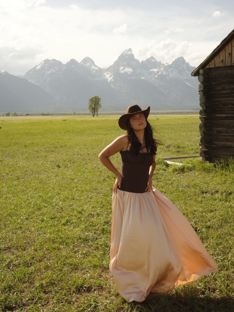 Woman wearing a brown corset-style bodice dress with a flowing peach satin skirt and a brown cowboy hat in a grassy field with mountains behind her