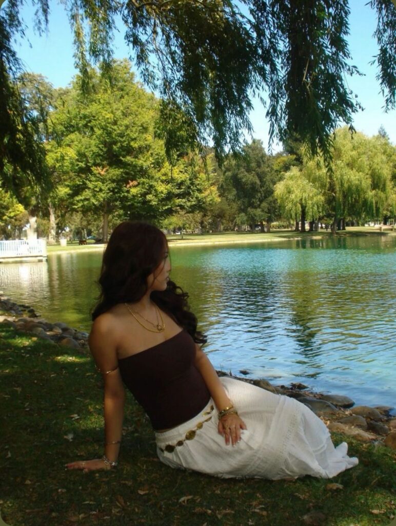 Woman wearing a brown tube top, flowy white maxi skirt, and gold jewelry sitting on the grass beside a calm lake