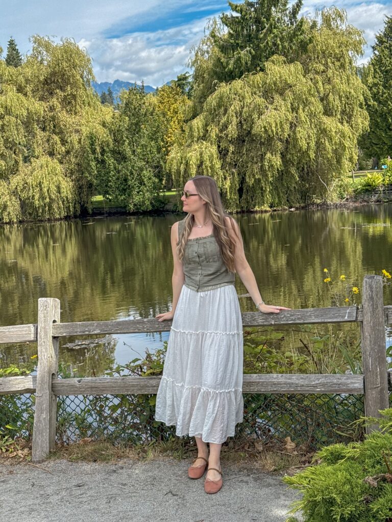 Woman wearing an olive green button-front tank top and white tiered midi skirt with ballet flats standing at a wooden fence beside a pond