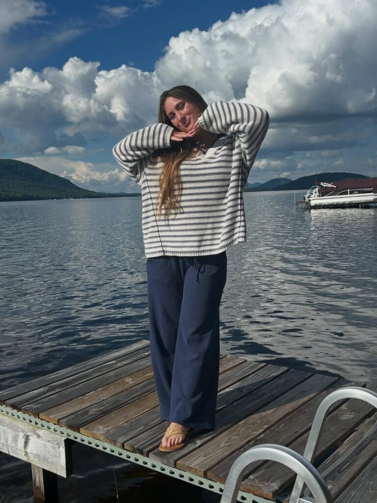 Woman wearing an oversized striped sweater, navy wide-leg pants, and sandals standing on a wooden dock over a lake