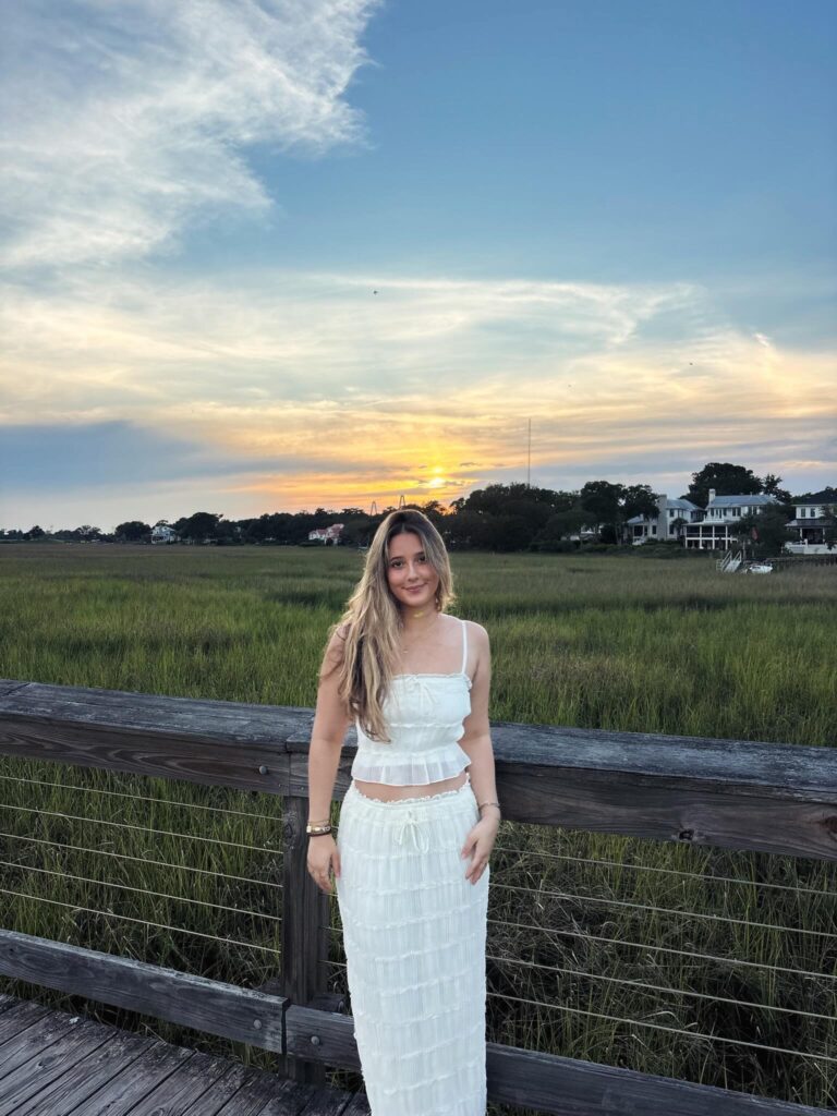 Woman wearing a white ruffled cami top and matching white maxi skirt standing on a boardwalk over tall grass at sunset