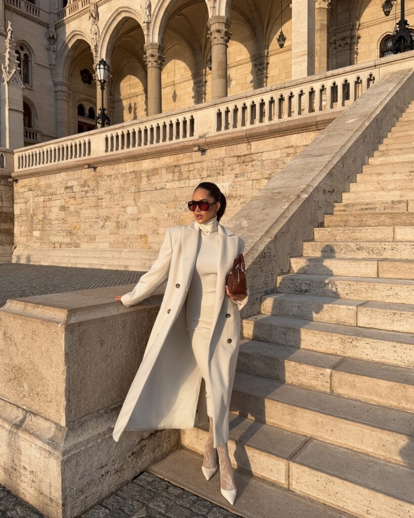 Woman on stone steps in Budapest wearing a long cream double-breasted coat over a matching cream knit turtleneck and skirt with white heels