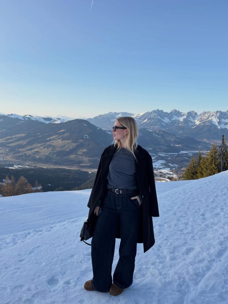 Woman standing on a snowy Alpine viewpoint in a black wool coat, grey knit top, dark wide-leg jeans and brown Ugg boots