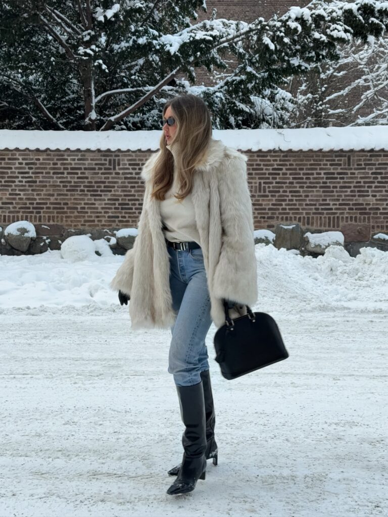 Woman walking through snow in a cream faux fur coat over a white knit, light-wash jeans, black knee-high boots and a black handbag