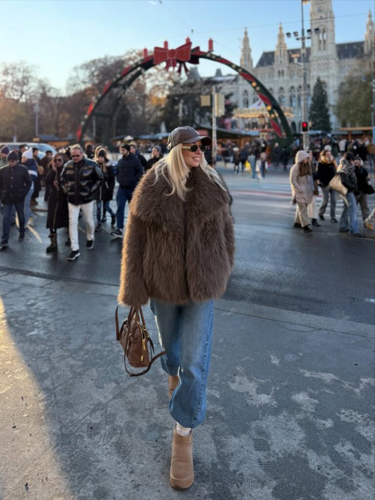 Woman crossing a square at a Vienna Christmas market in a brown faux fur jacket, straight-leg jeans, tan Ugg boots and a brown leather cap