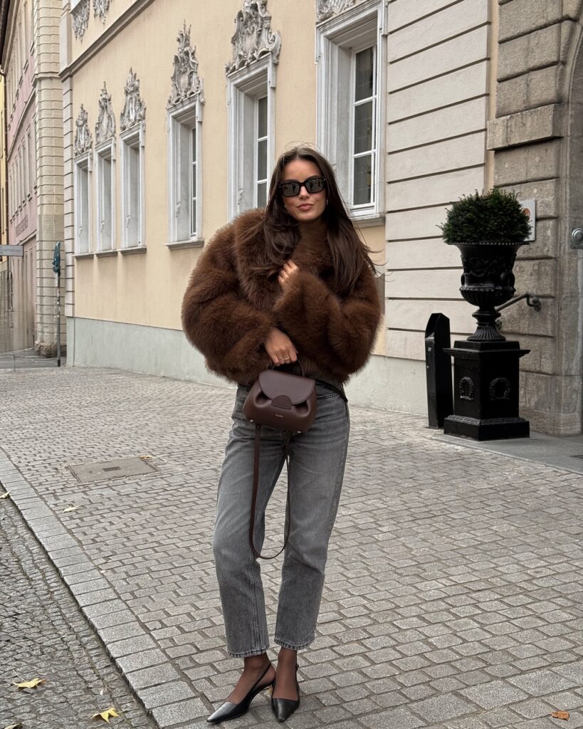 Woman on a cobbled European street in a cropped brown faux fur jacket with grey straight-leg jeans, black slingback flats and a brown mini bag