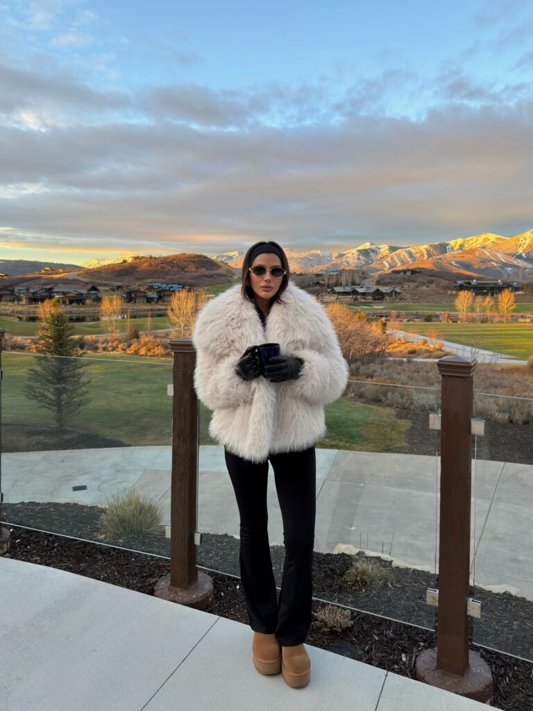 Woman holding a mug at sunrise in a cream faux fur coat with black flared trousers, tan platform boots, black gloves and a headband