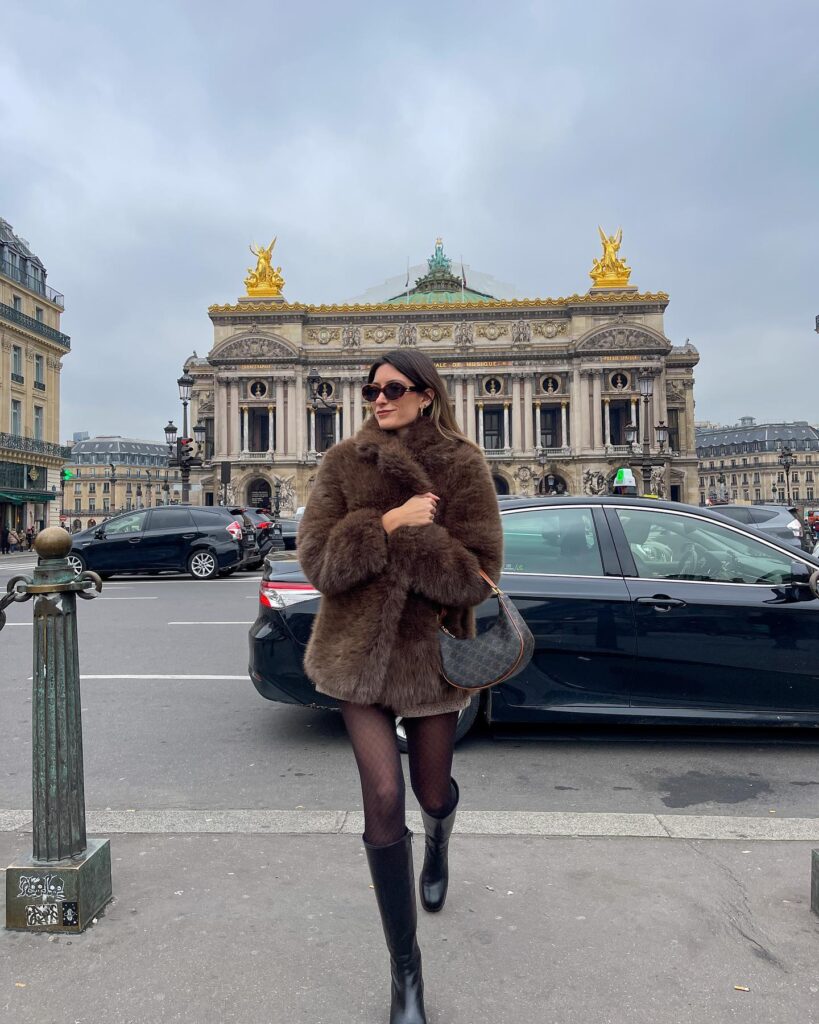 Woman in front of the Paris Opéra wearing a brown faux fur coat, sheer black tights, tall black riding boots and a monogram shoulder bag