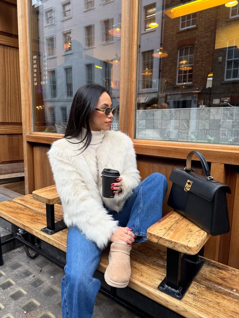 Woman sitting on a wooden bench outside a London coffee shop in a white faux fur jacket, cream turtleneck, blue jeans and sand Ugg boots