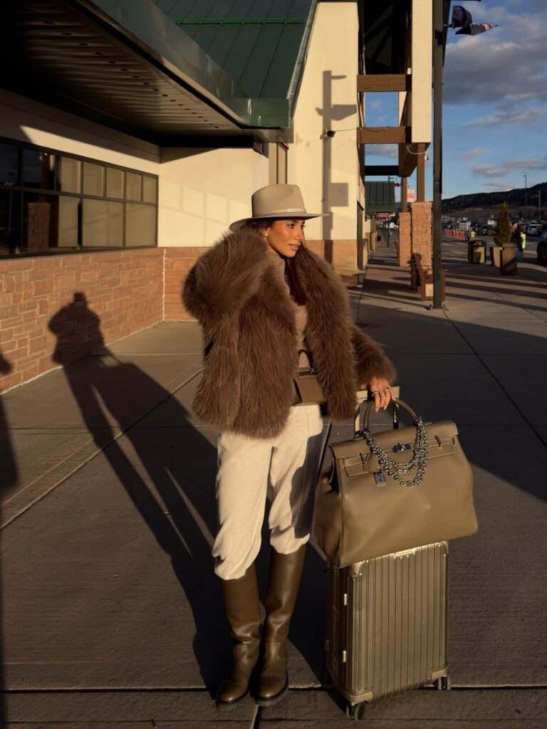 Woman with a suitcase and large tote wearing a brown faux fur coat, taupe fedora, cream trousers and tall olive leather boots