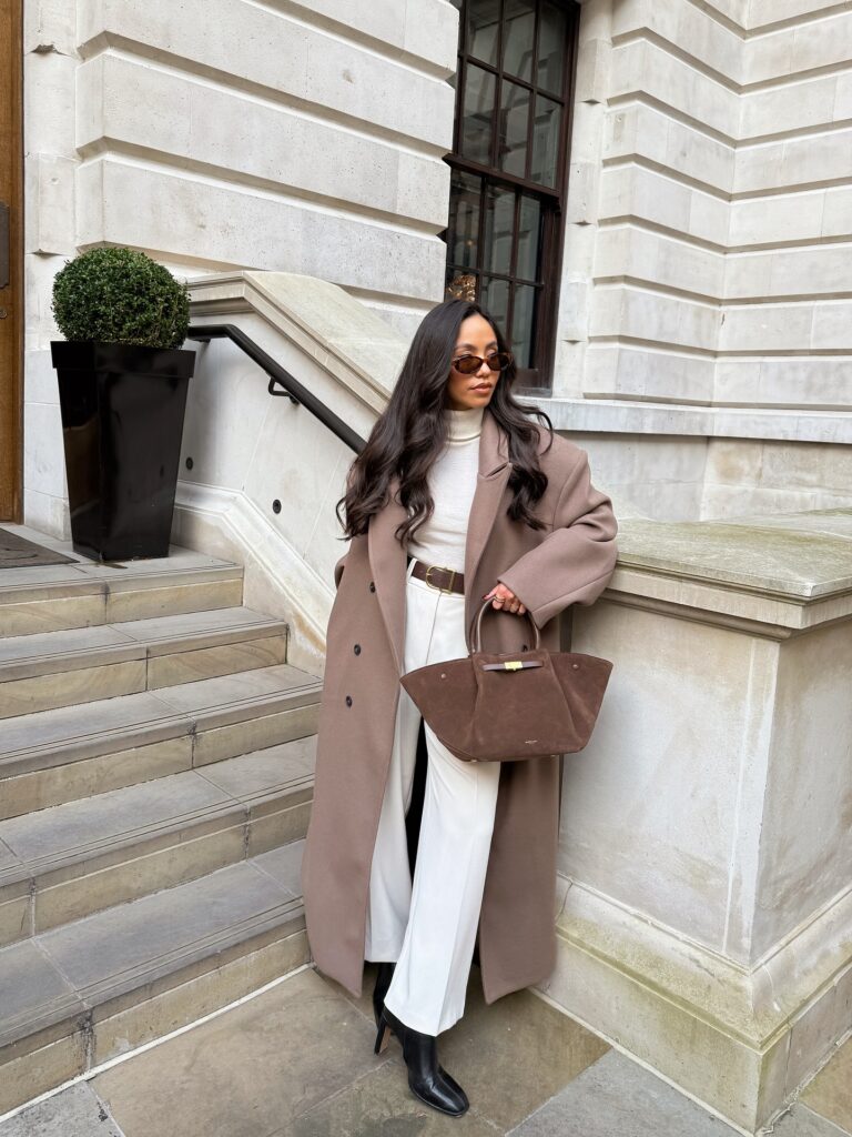 Woman in an oversized mocha brown wool coat with a cream turtleneck, wide-leg white trousers, brown suede tote and black boots