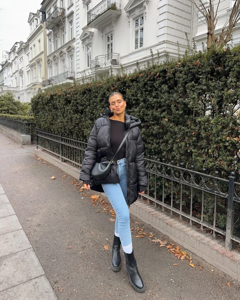 Woman on a Hamburg street in an oversized black hooded puffer jacket with a black top, light-wash skinny jeans, white socks and black Chelsea boots