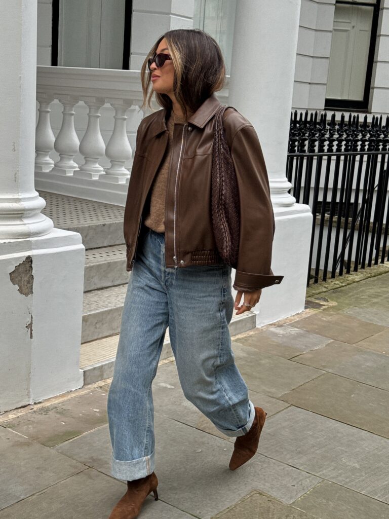 Woman walking a London street in a brown leather bomber jacket over a camel knit with cuffed baggy jeans and brown suede ankle boots