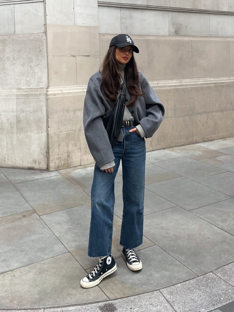 Woman in a grey wool bomber jacket over a grey turtleneck with dark cropped wide-leg jeans, black high-top Converse and a black cap