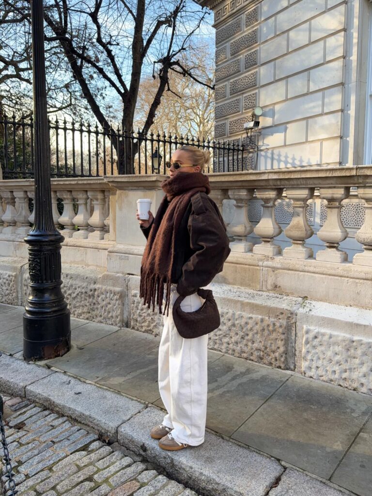 Woman on a London pavement in a brown suede jacket with an oversized chocolate fringed scarf, white wide-leg jeans and taupe trainers