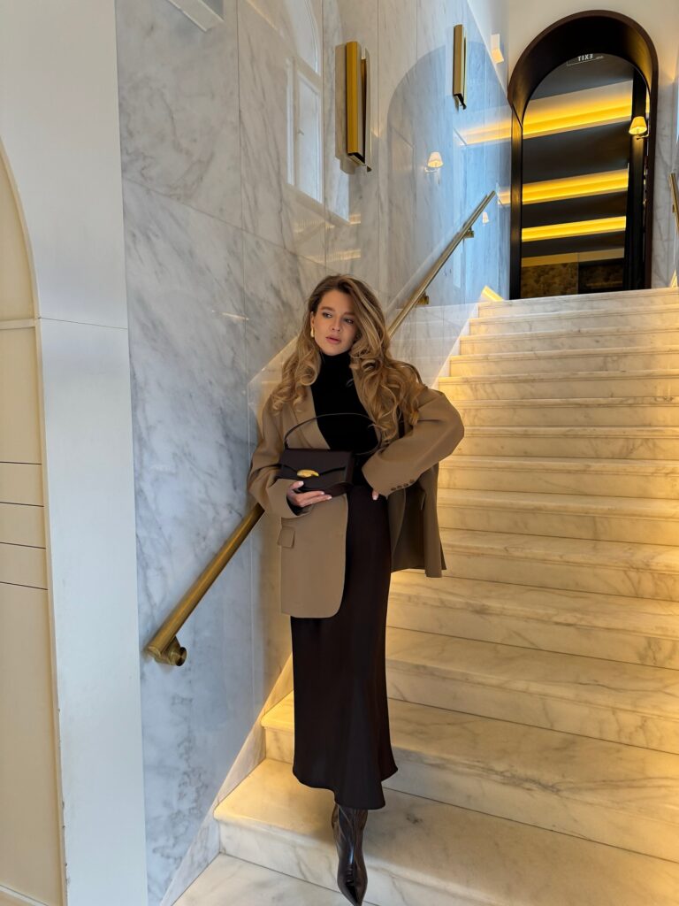 Woman on a marble hotel staircase in an oversized camel blazer over a black turtleneck with a black satin midi skirt and black heeled boots