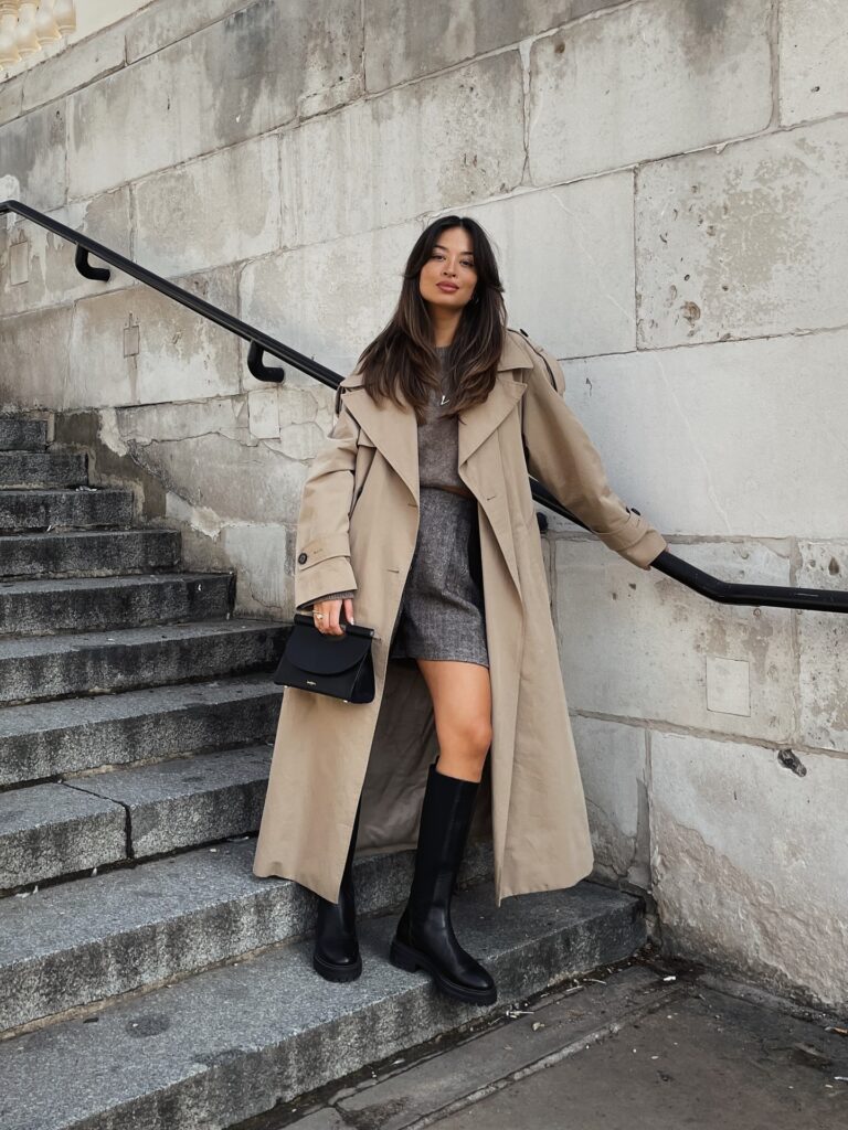 Woman on stone steps in a long beige trench coat over a grey knit and grey herringbone mini skirt with tall black boots