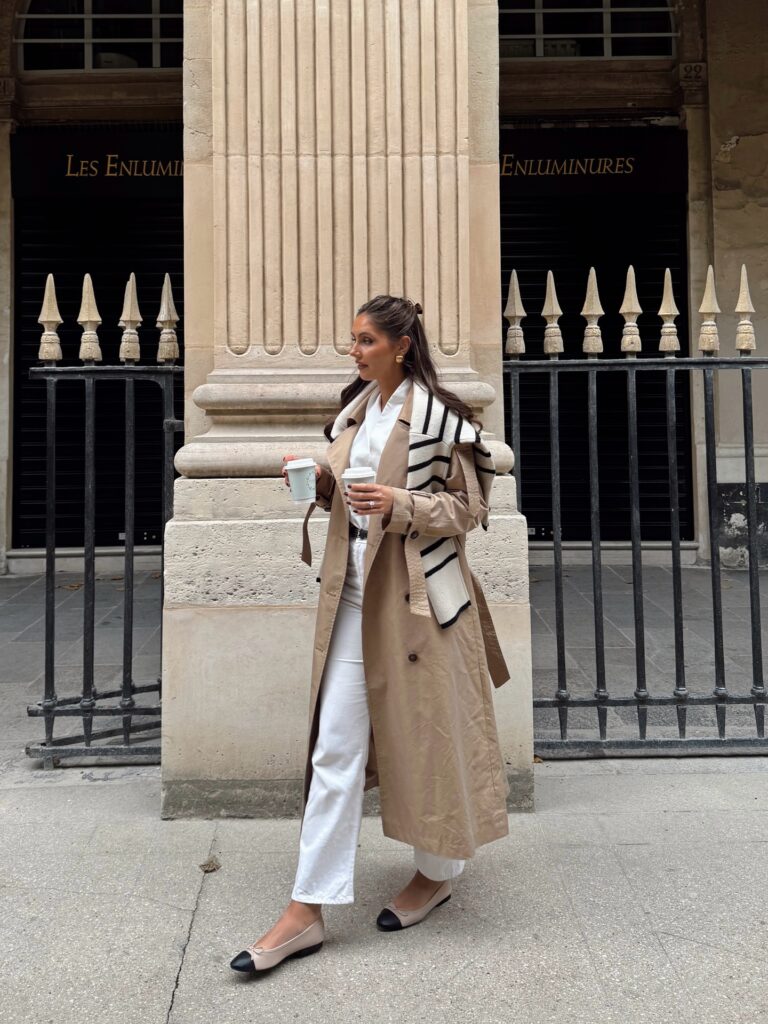Woman in Paris holding two coffees in a beige trench coat with a cream and navy striped scarf, white shirt, white jeans and two-tone ballet flats