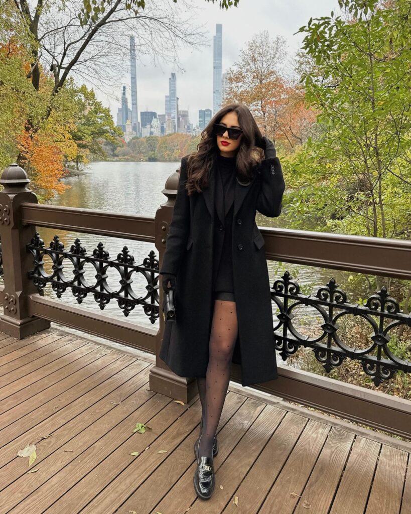 Woman on a park bridge in a long black coat over a black blazer and shorts with sheer polka dot tights, black chunky loafers and gloves
