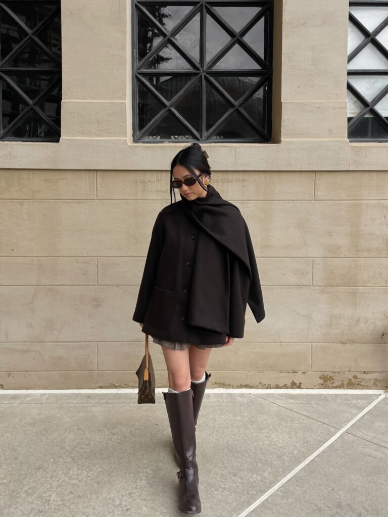 Woman against a stone wall in an oversized black wool jacket with a matching draped scarf, grey plaid mini skirt, knee socks and brown riding boots