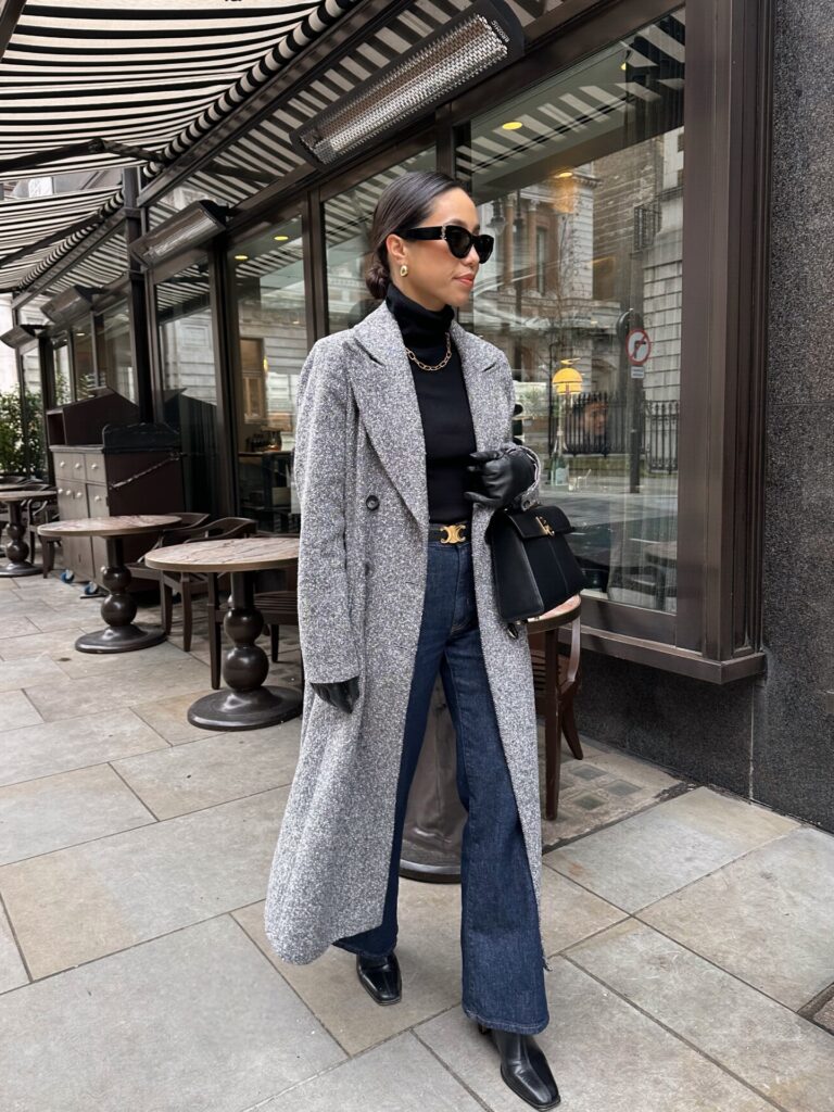 Woman in a long grey herringbone coat with a black turtleneck, gold chain necklace, dark flared jeans, leather gloves and a black top-handle bag